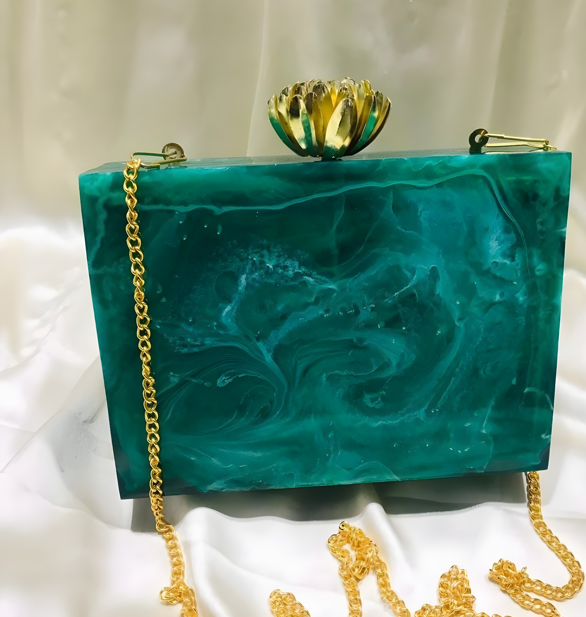 A stylish teal and gold handbag with a textured surface and a decorative top, resting on a light-colored fabric.