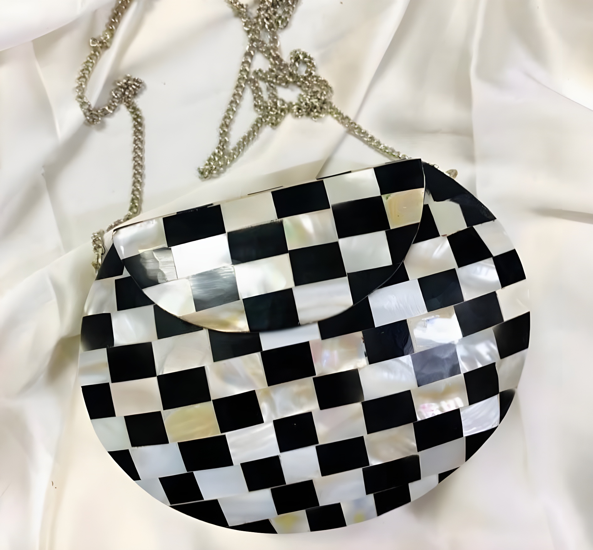 A stylish black and white checkered handbag with a curved shape and silver chain strap, resting on a light-colored fabric background.